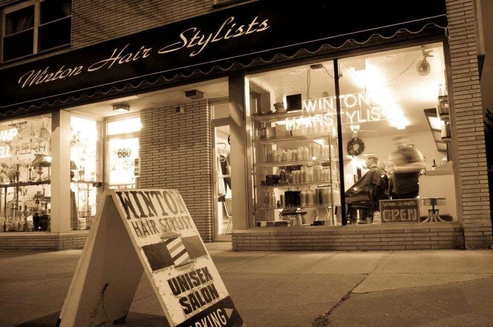 The shop at night: Winton Hair Stylists in script on the awning, the front window lit up, and the sidewalk sign out front